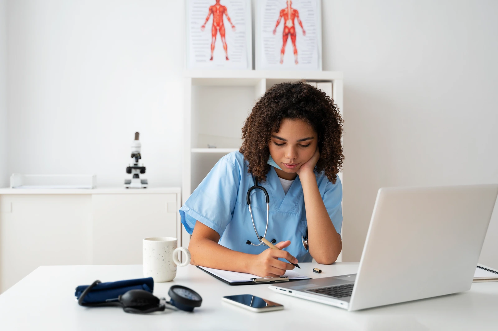 Tired nursing student in scrubs at a desk with a laptop and notes